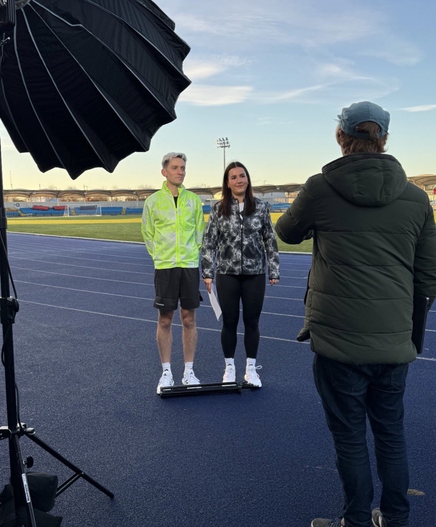 Two running friendly - man and a woman - standing on an athletics track, with cameras in front of them. They are about to film some content.